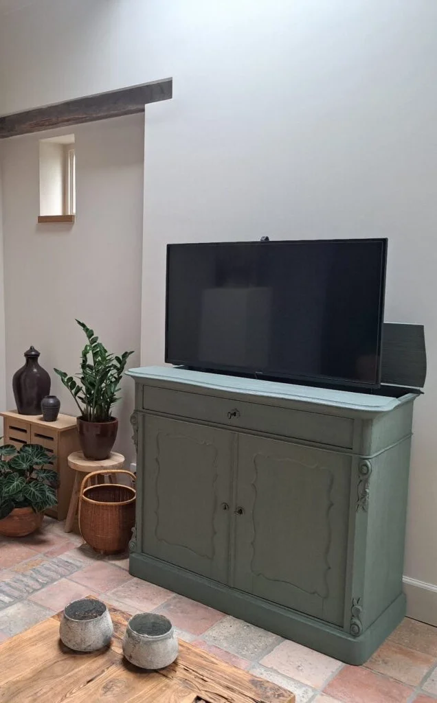 Groene vintage kast met een TV lift bovenop, omgeven door planten in een lichte ruimte met stenen vloer en houten bijzettafel.
