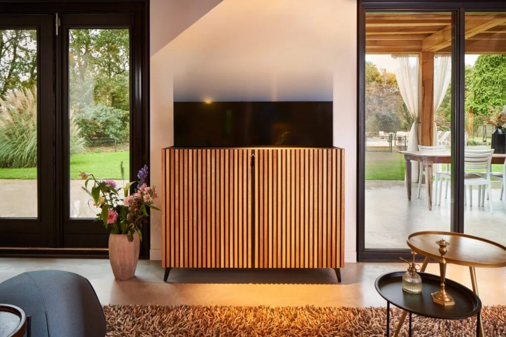 Modern houten TV-meubel met verticale lijnen, omringd door grote ramen en een gezellige zithoek met bloemen en tafeltjes.