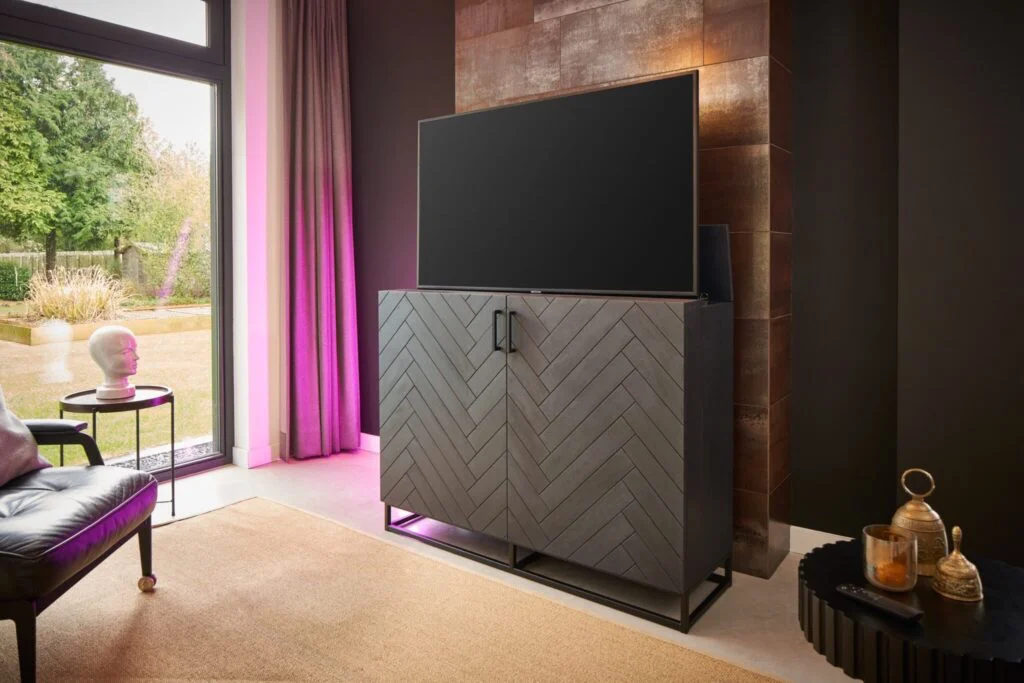 Donkergrijs kastje met visgraatmotief en TV. Modern interieur met leren stoel, bijzettafel en uitzicht op de tuin.