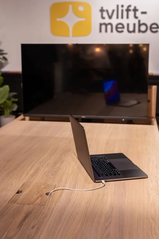 Laptop op houten tafel met moderne, minimalistische uitstraling, TV lift meubel op achtergrond, groene plant aan de zijkant.