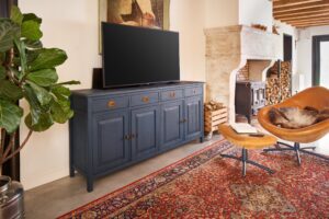 Donkerblauw houten dressoir met tv, vintage vloerkleed, leren stoel en open haard. Rustieke en stijlvolle woonkamerinrichting.