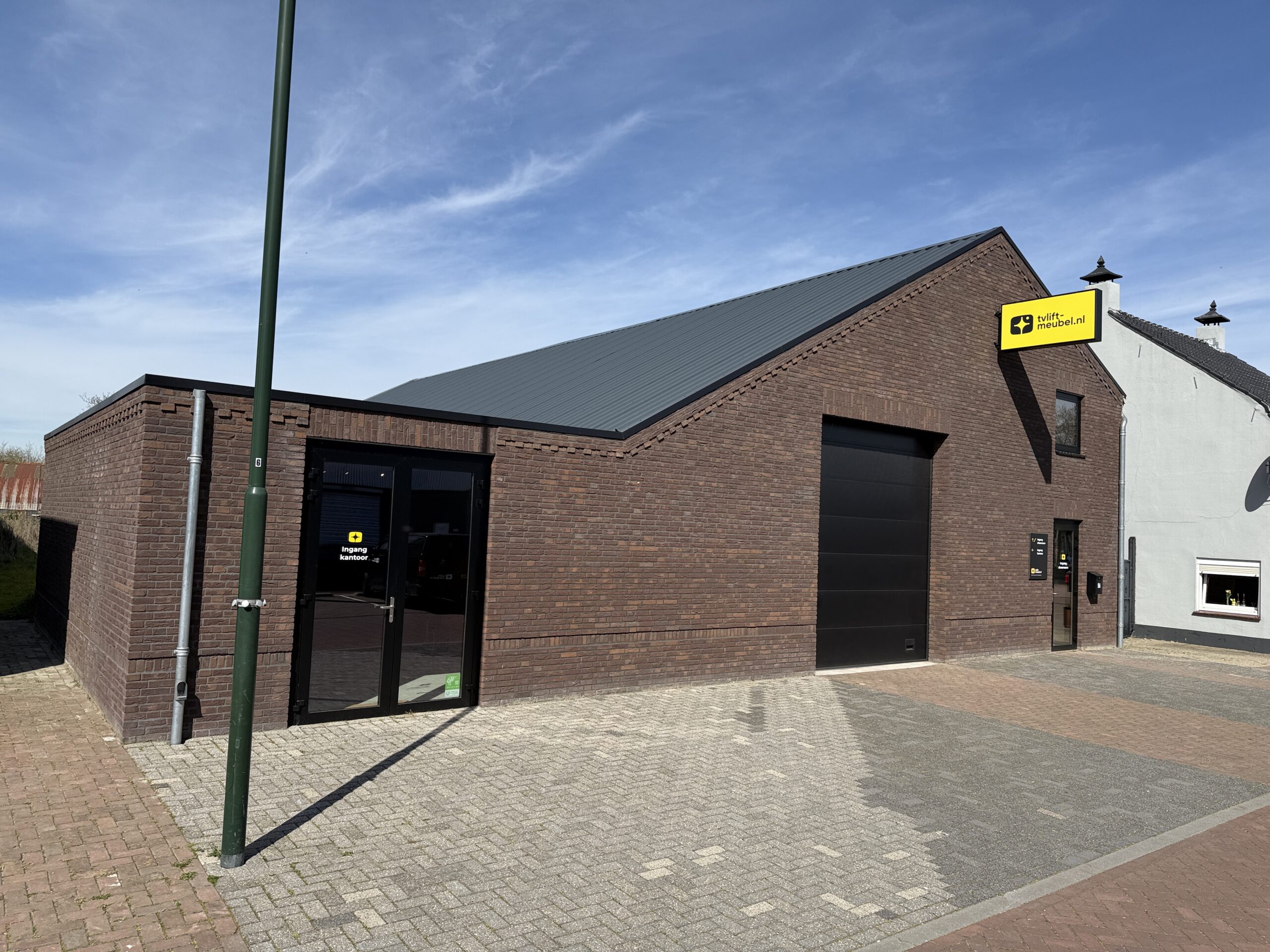 Bakstenen bedrijfsgebouw met schuine dakconstructie en glazen entree, voorzien van een helder geel bedrijfslogo bord in een rustige straat.